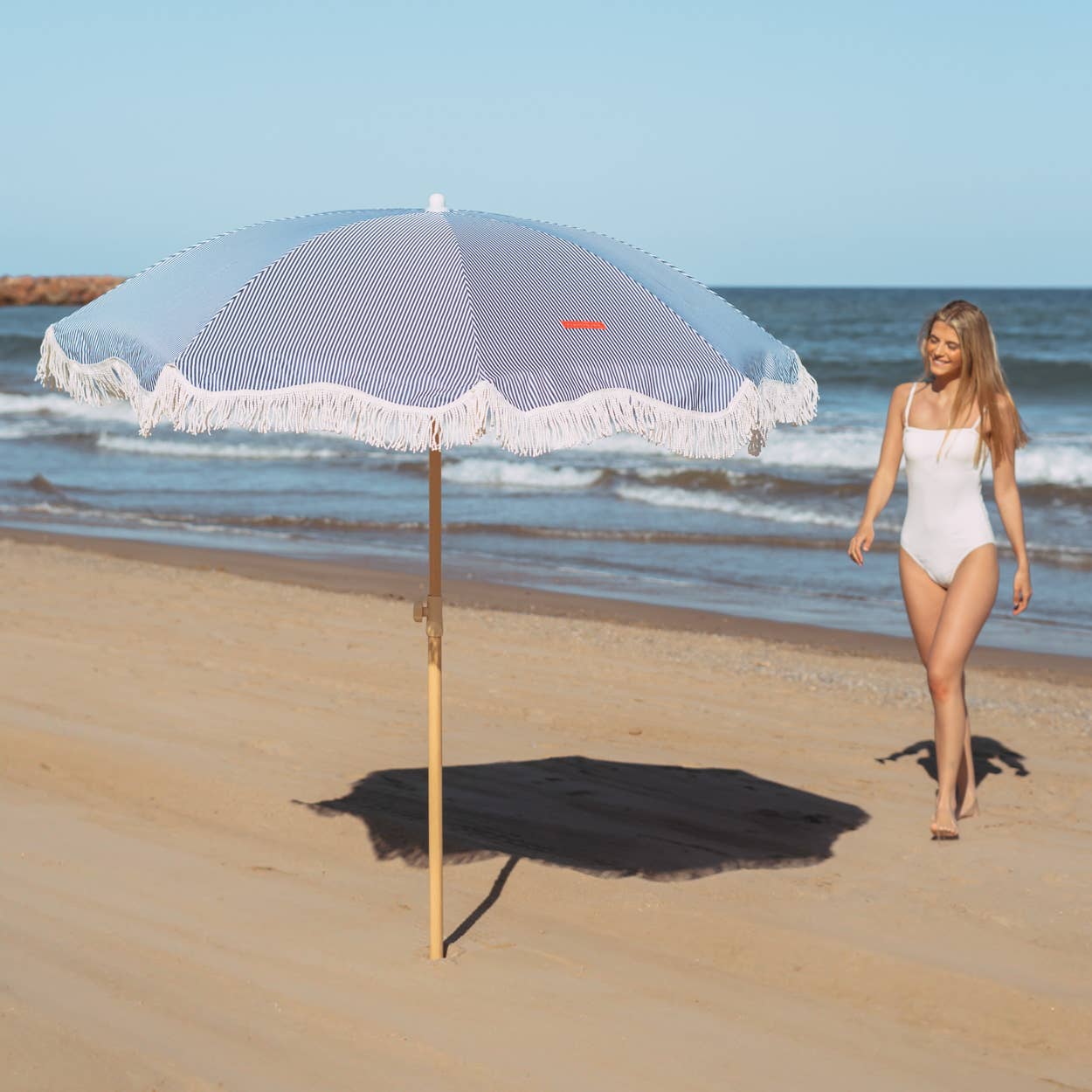 En kvinne går på stranden ved siden av den oppsatte blå-stripete strandparasollen CAPRI, som gir skygge og beskyttelse mot solen.