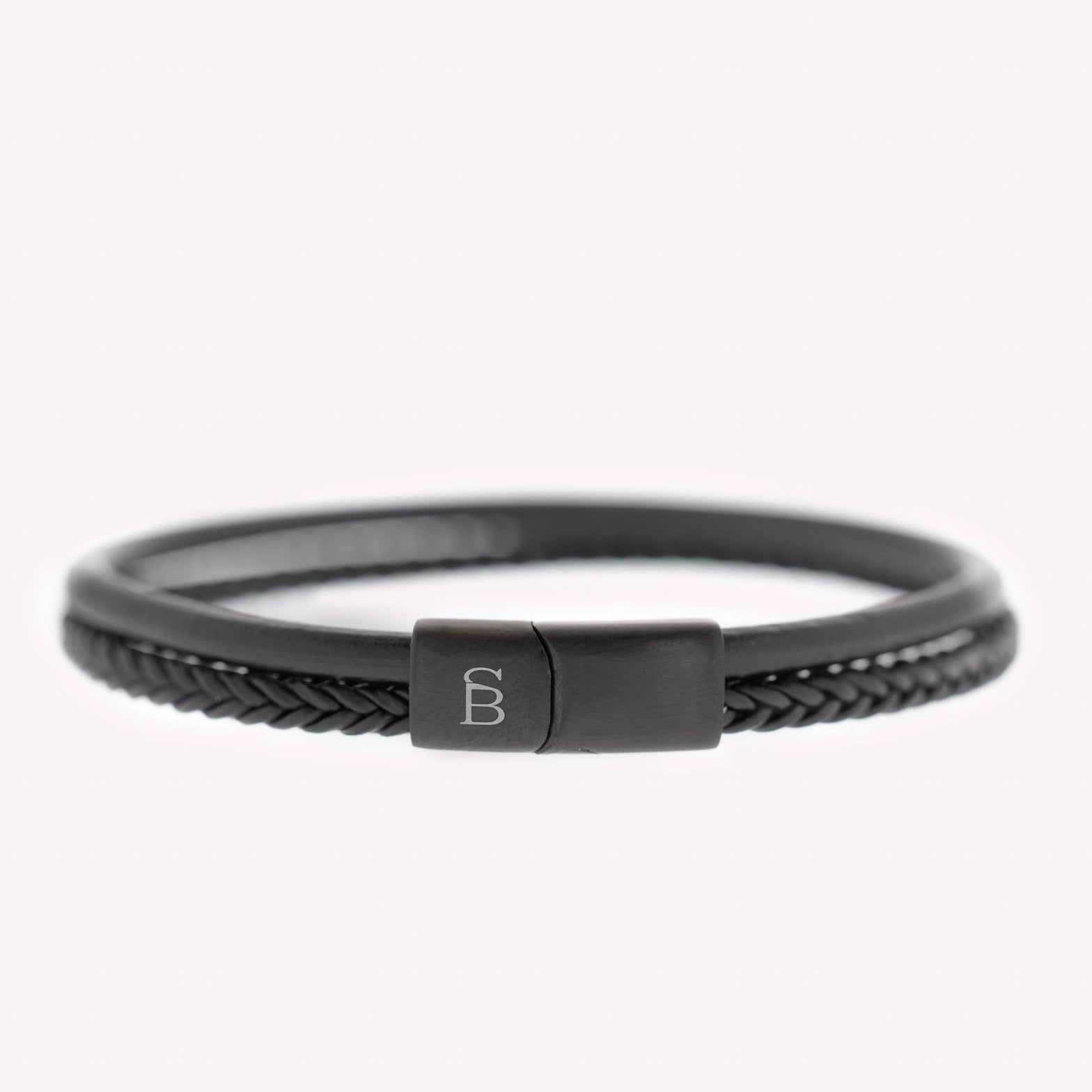 Et minimalistisk svart lærarmbånd fra Steel & Barnett, med en matt, svart magnetisk lås gravert med initialene "SB." Armbåndet ligger mot en lys, nøytral bakgrunn som fremhever det elegante designet.