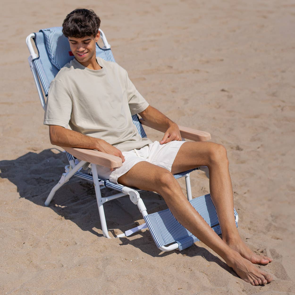 En ung mann sitter avslappet i en blå-hvit stripete strandstol på stranden, med armlener i tre og en justerbar fotstøtte.