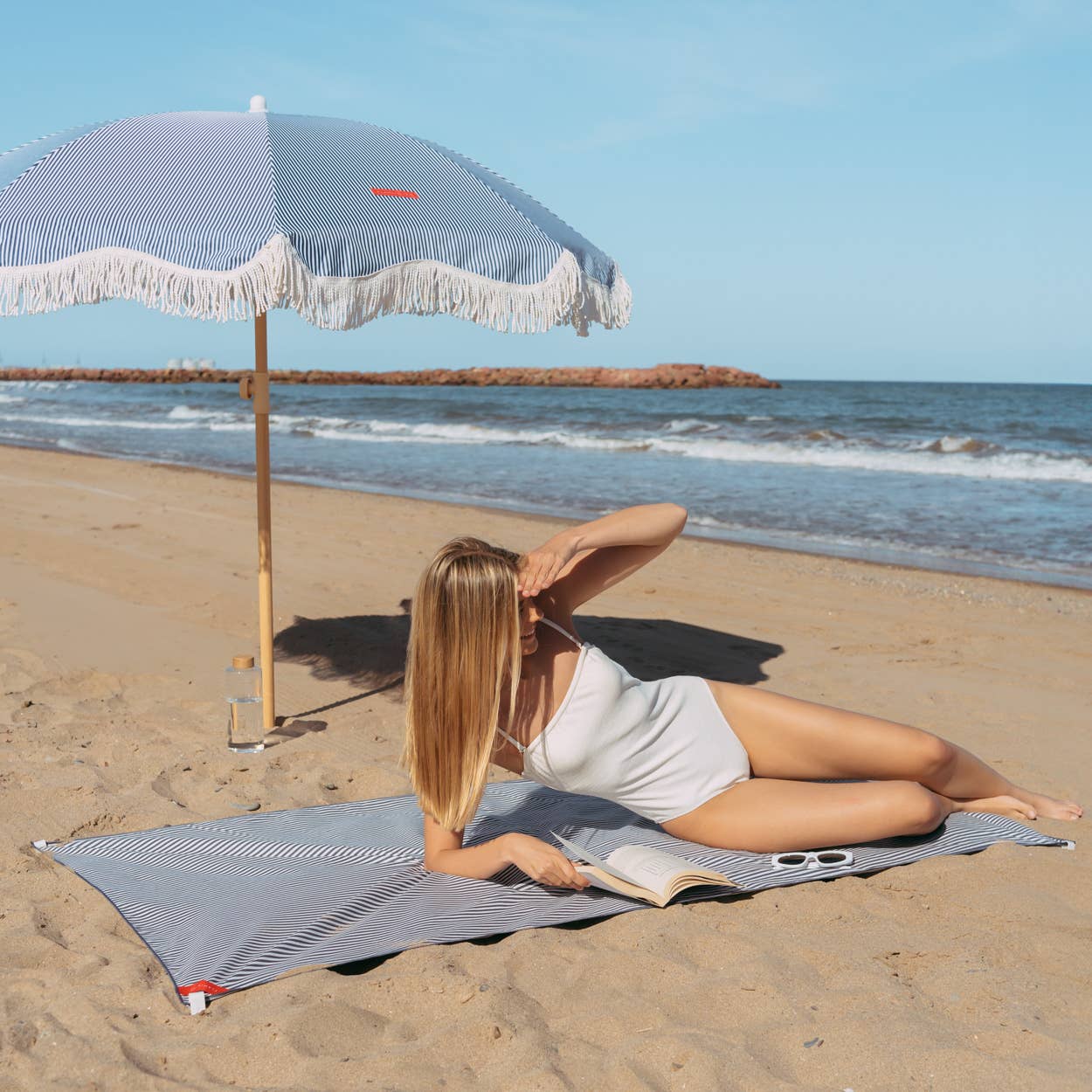 En kvinne slapper av på stranden under den blå-stripete strandparasollen CAPRI, som gir skygge og stil med sine dekorative frynser.