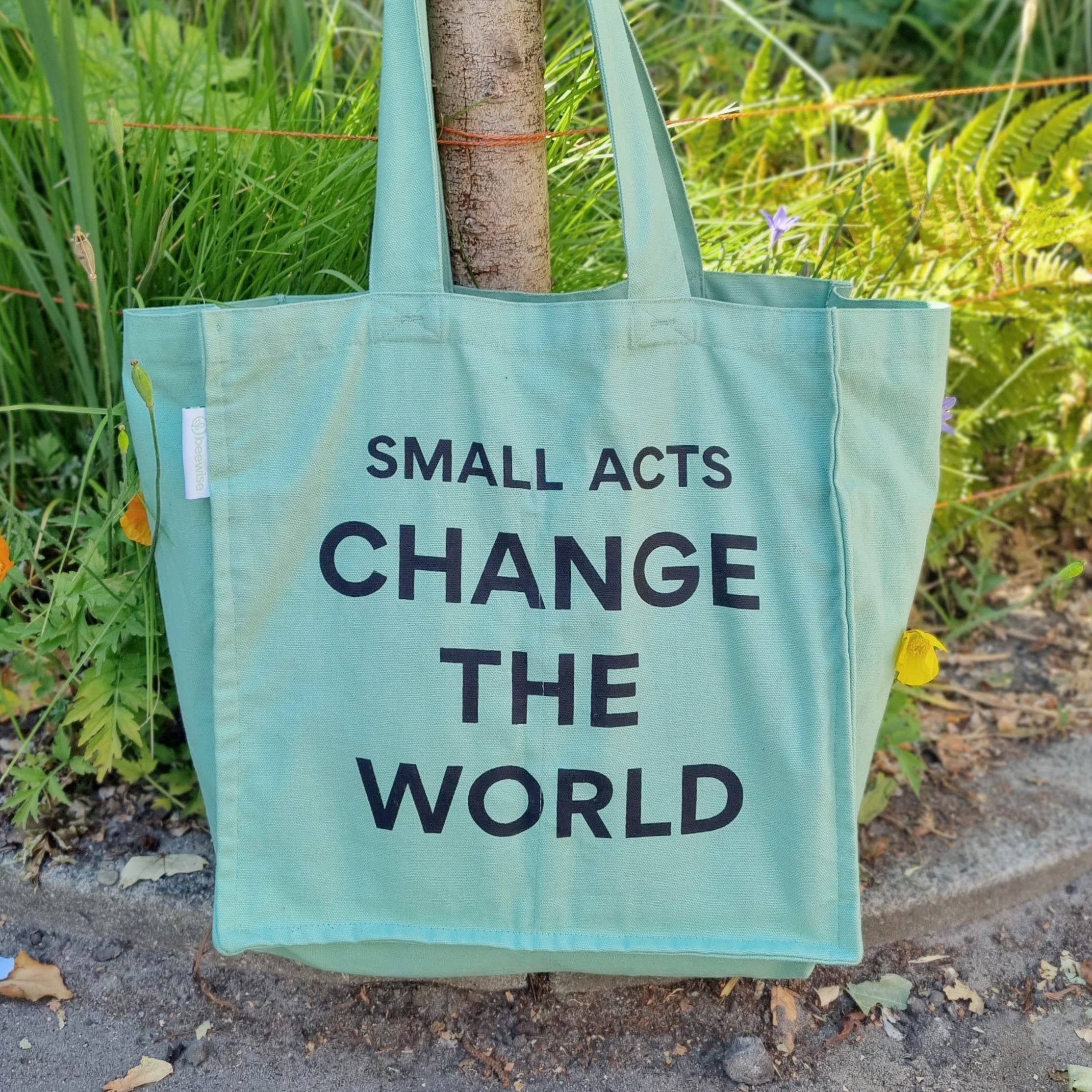 Mintgrønn handlenett i økologisk bomull med teksten 'Small acts change the world', hengende på et tre.