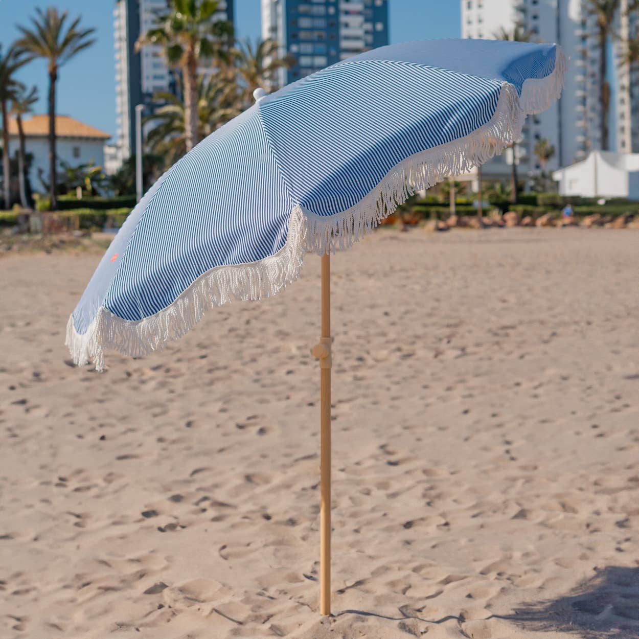 Blå-stripete strandparasoll CAPRI står oppreist på stranden, med tofarget polyesterstoff og frynser som gir et elegant utseende.