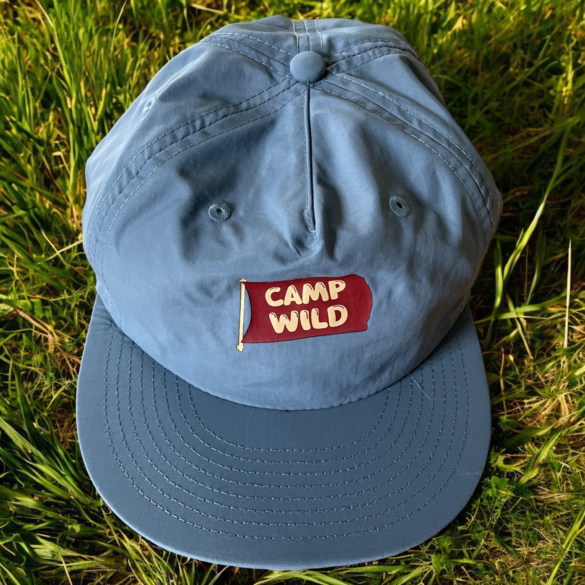 Glacier blå Camp Wild Caps med rød flagglogo, plassert på gress. Capsen er laget av resirkulert nylonstoff og er lett og hurtigtørkende.