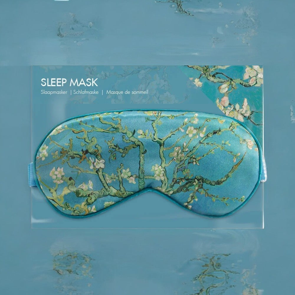 Sovemasken med "Almond Blossom"-motivet er plassert i sin emballasje, som har en lyseblå bakgrunn med teksten "SLEEP MASK" i hvitt. Bakgrunnen inkluderer også elementer av kunstverket.