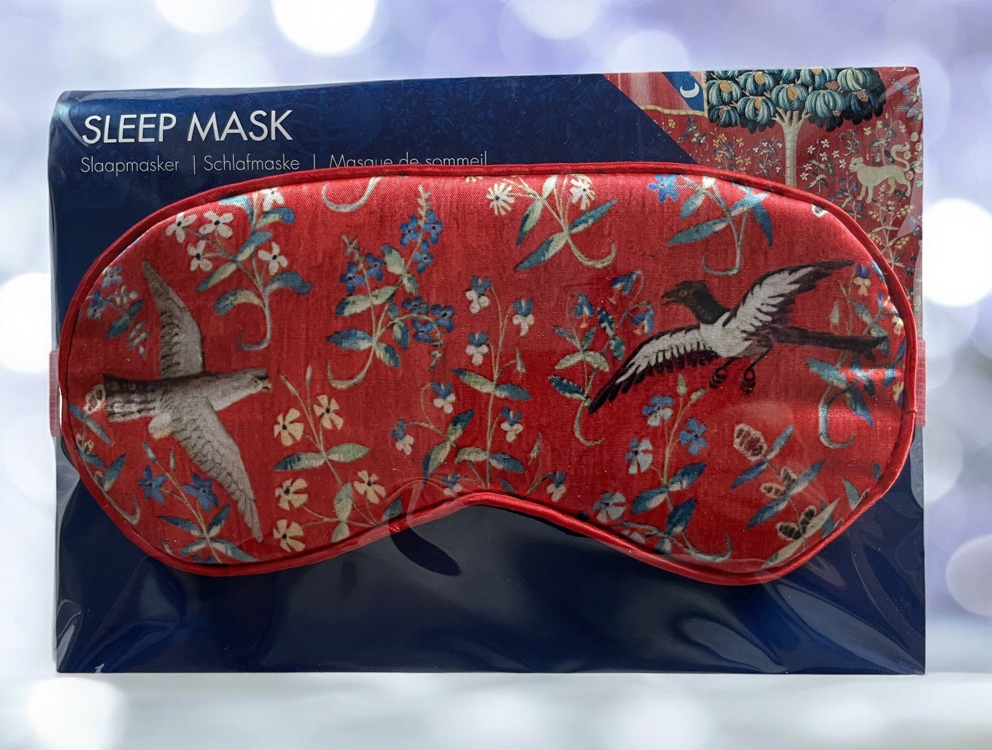 Den røde sovemasken med fuglemotiv er plassert i sin emballasje. Bakgrunnen har et utsnitt fra "The Lady and the Unicorn" tapetet, og teksten "SLEEP MASK" står øverst på emballasjen.