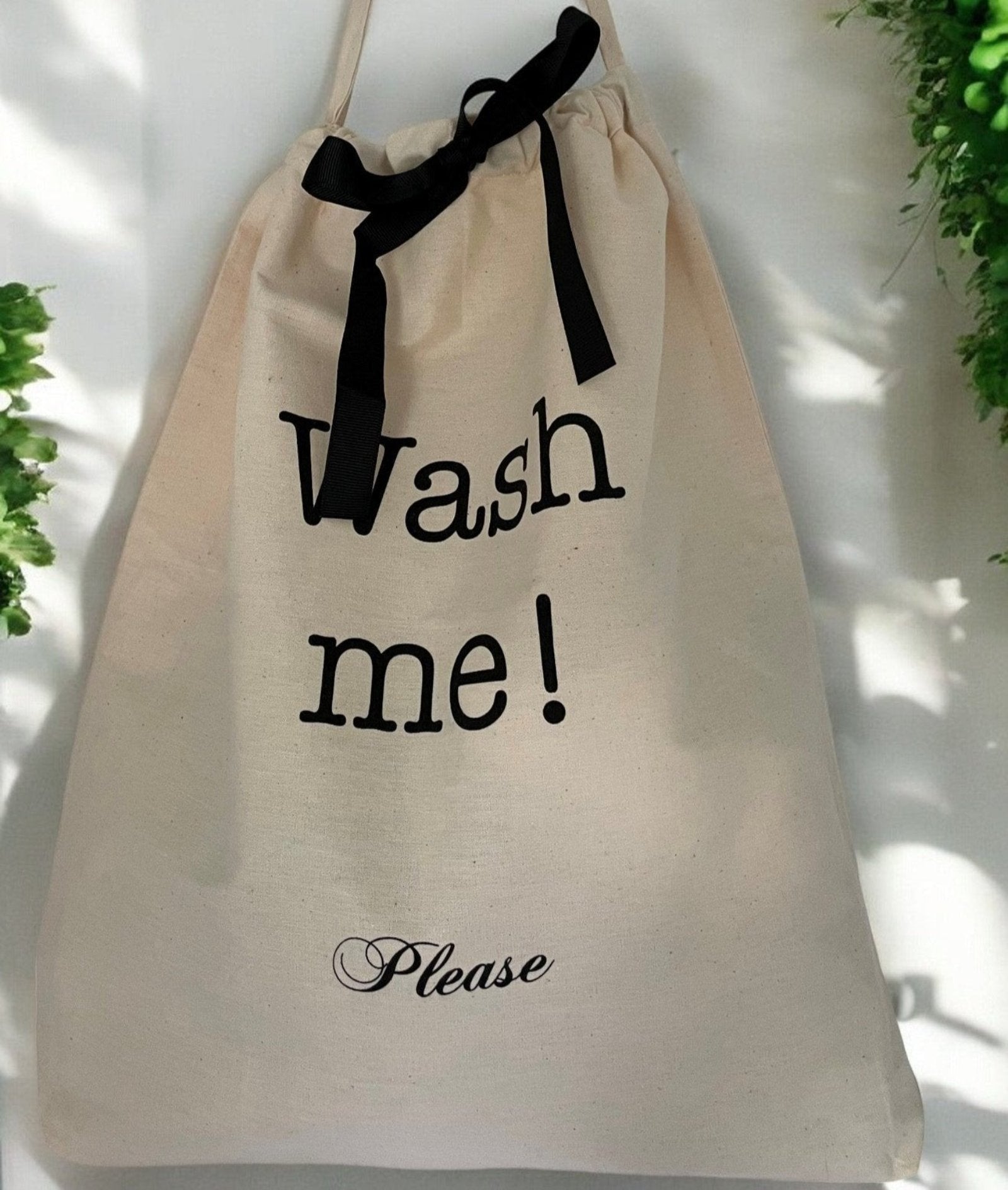 En Bag-All pose med teksten 'Wash me!' og 'Please' i sort trykk, hengt på en hvit vegg med grønne planter i bakgrunnen. Posen er lukket med et sort bånd."