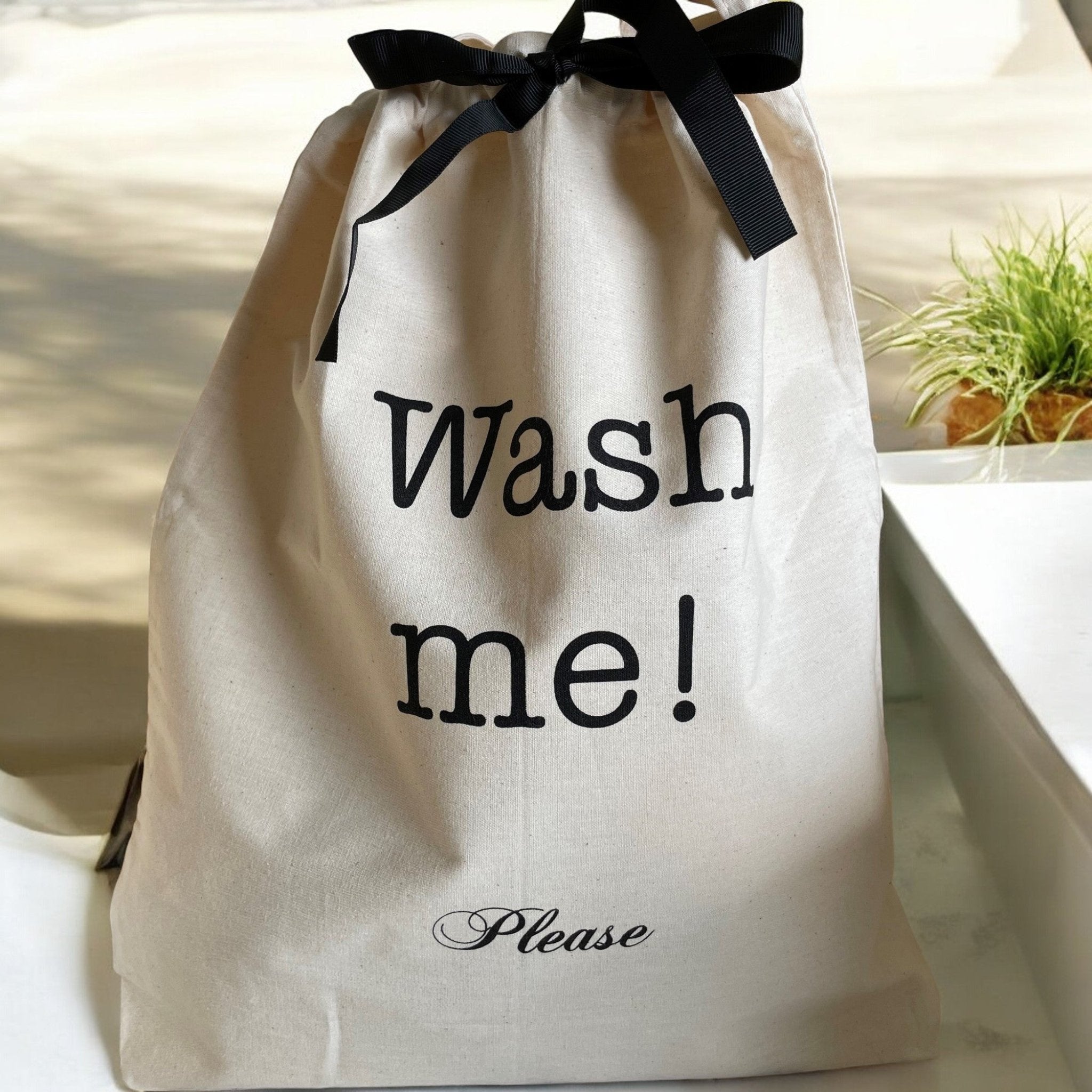 En Bag-All pose med teksten 'Wash me!' og 'Please' i sort trykk, plassert på en lys overflate med en grønn plante i bakgrunnen. Posen er lukket med et sort bånd.